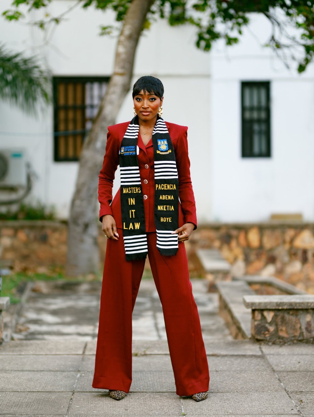 Woman in a red blazer and trousers wearing a graduation stole outdoors.