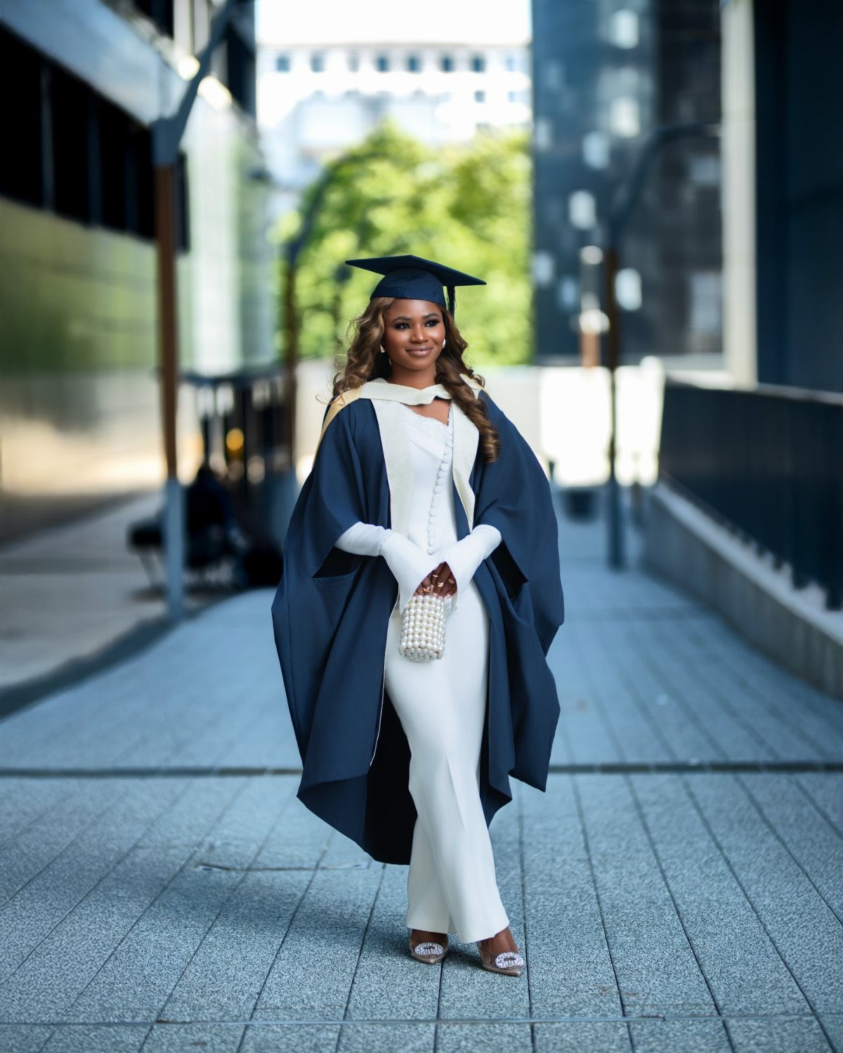 Woman in white jumpsuit and navy graduation gown posing on a city street