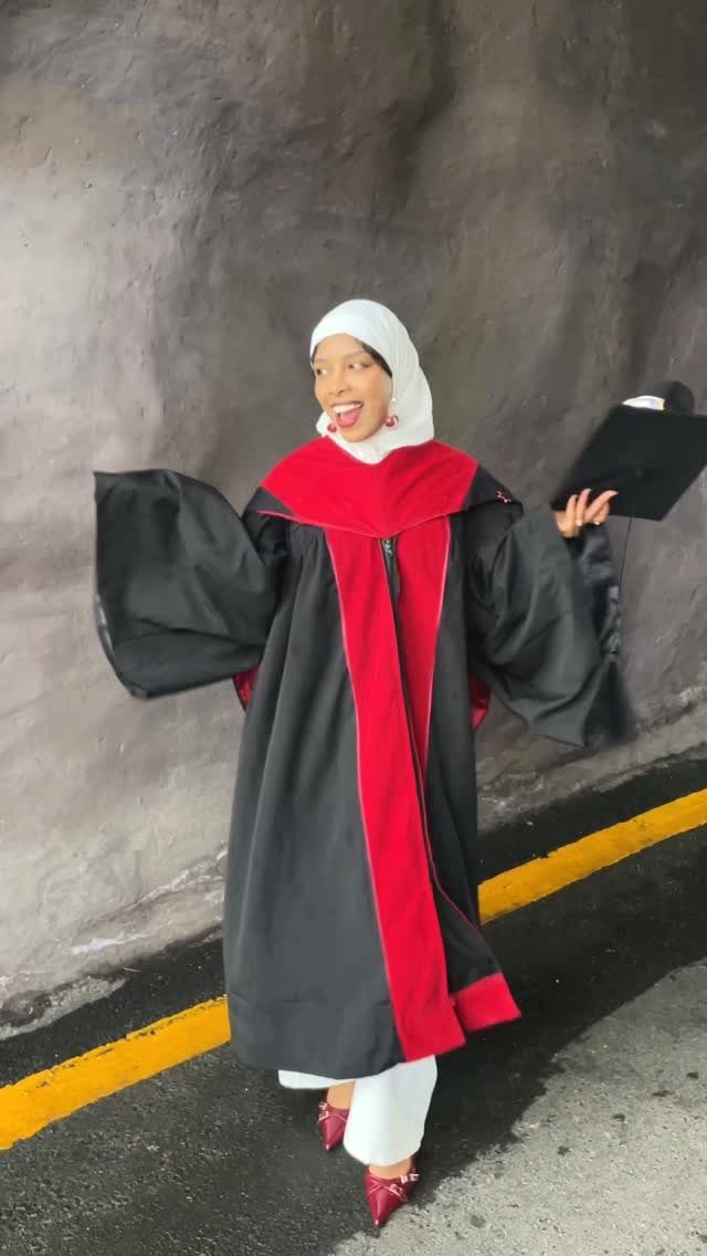 Woman in white hijab and black and red graduation gown posing outdoors.