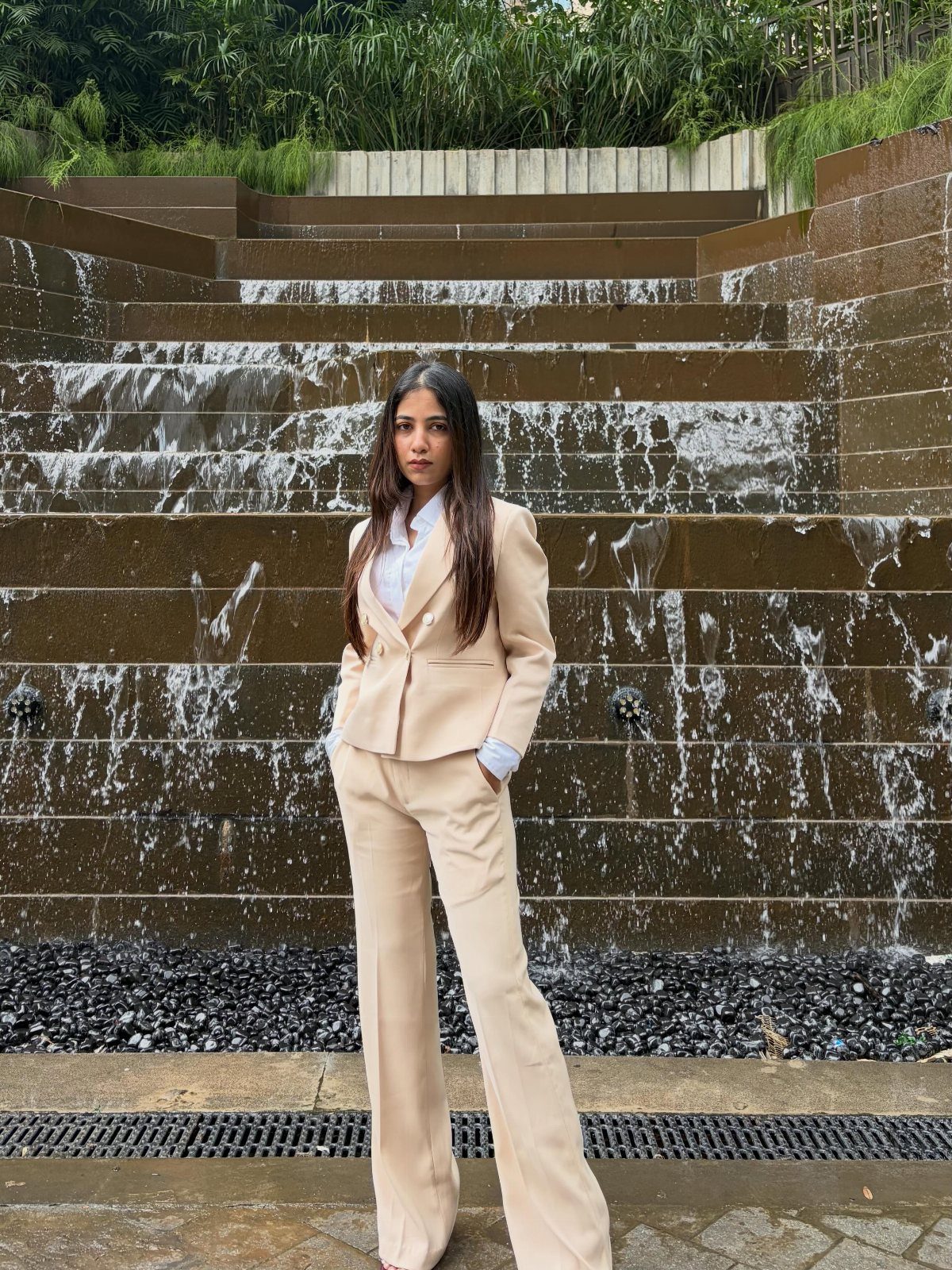 Woman in a beige blazer and matching trousers posing before a stone waterfall.