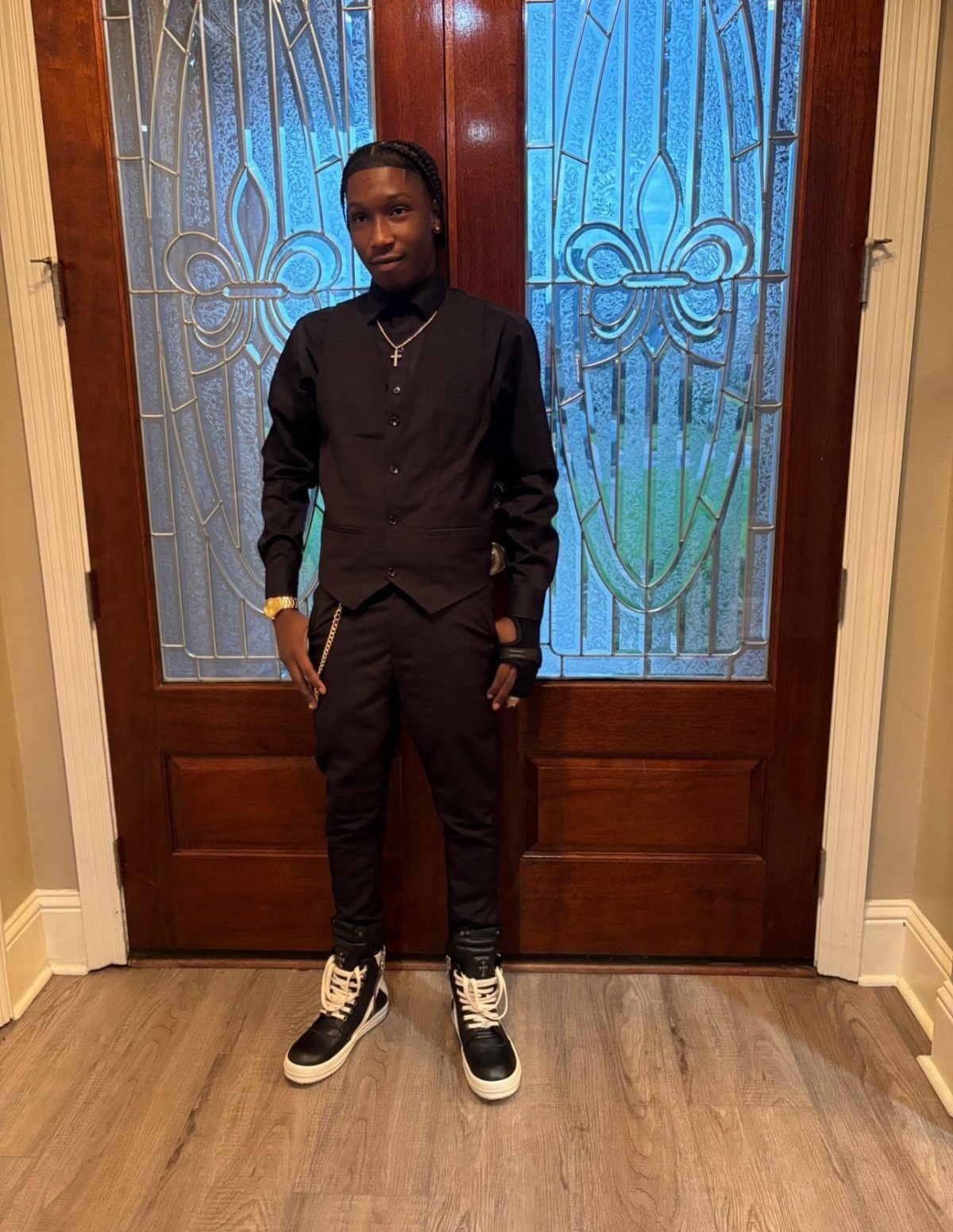 Young man wearing an all-black dressy outfit and sneakers standing before wooden doors.