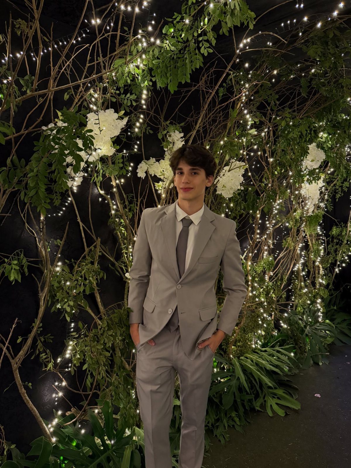 Young man in a light gray suit and tie posing before floral lights.