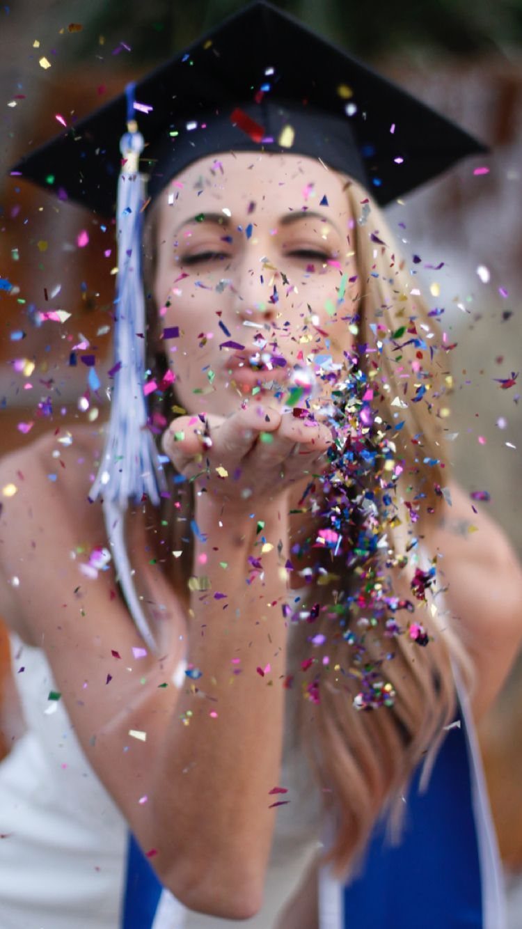 Graduate in cap and gown blowing colorful confetti toward the camera outdoors