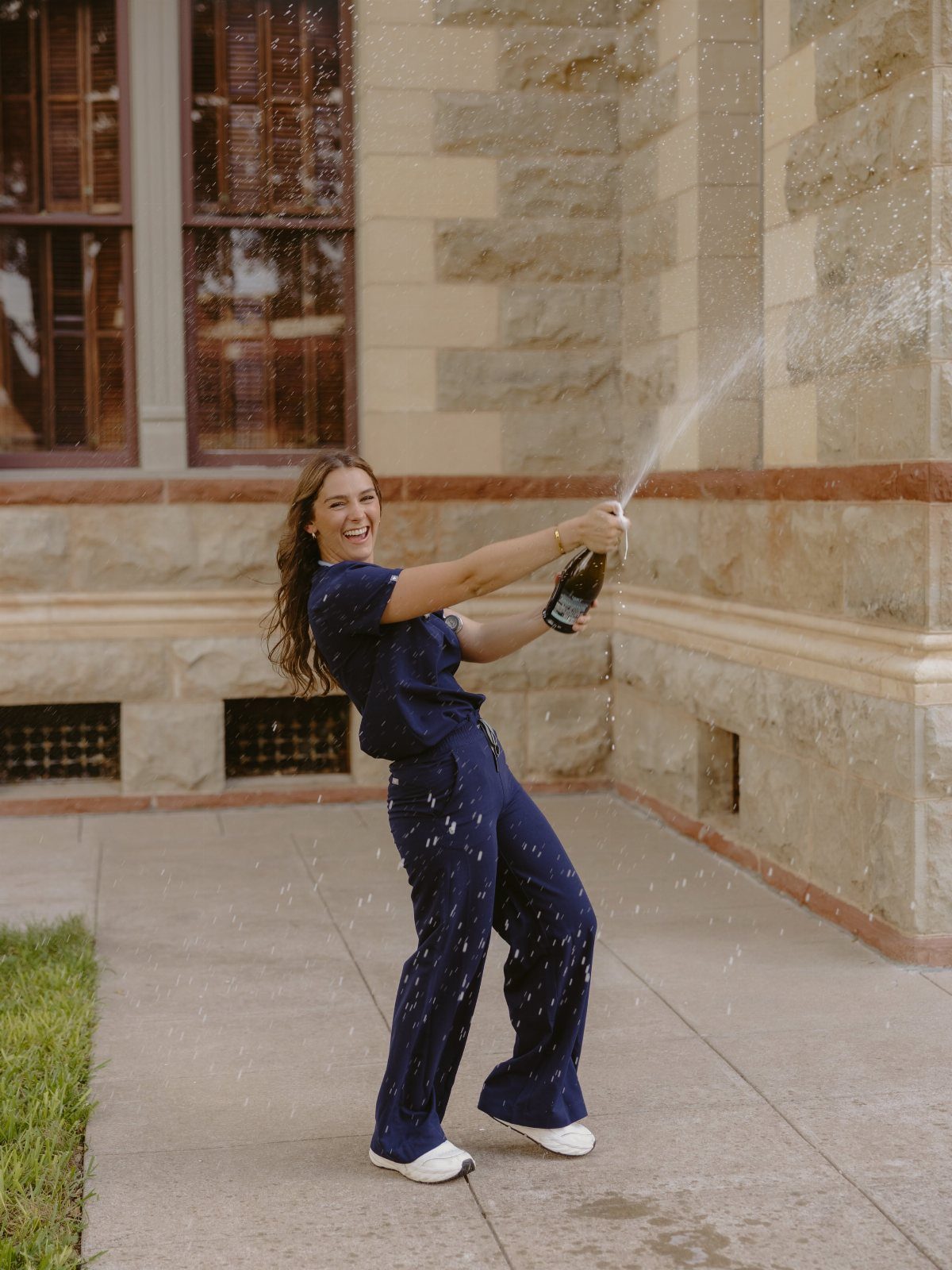 Woman in navy scrubs smiling while popping a bottle of champagne outdoors.