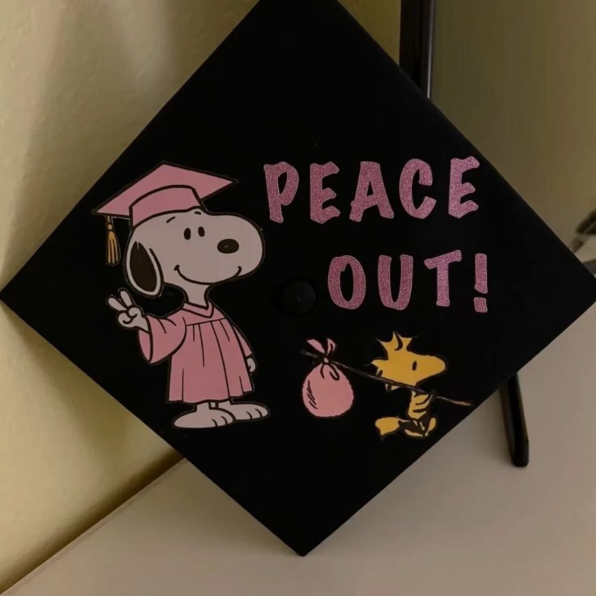 Black graduation cap decorated with pink Snoopy and Woodstock and text Peace Out