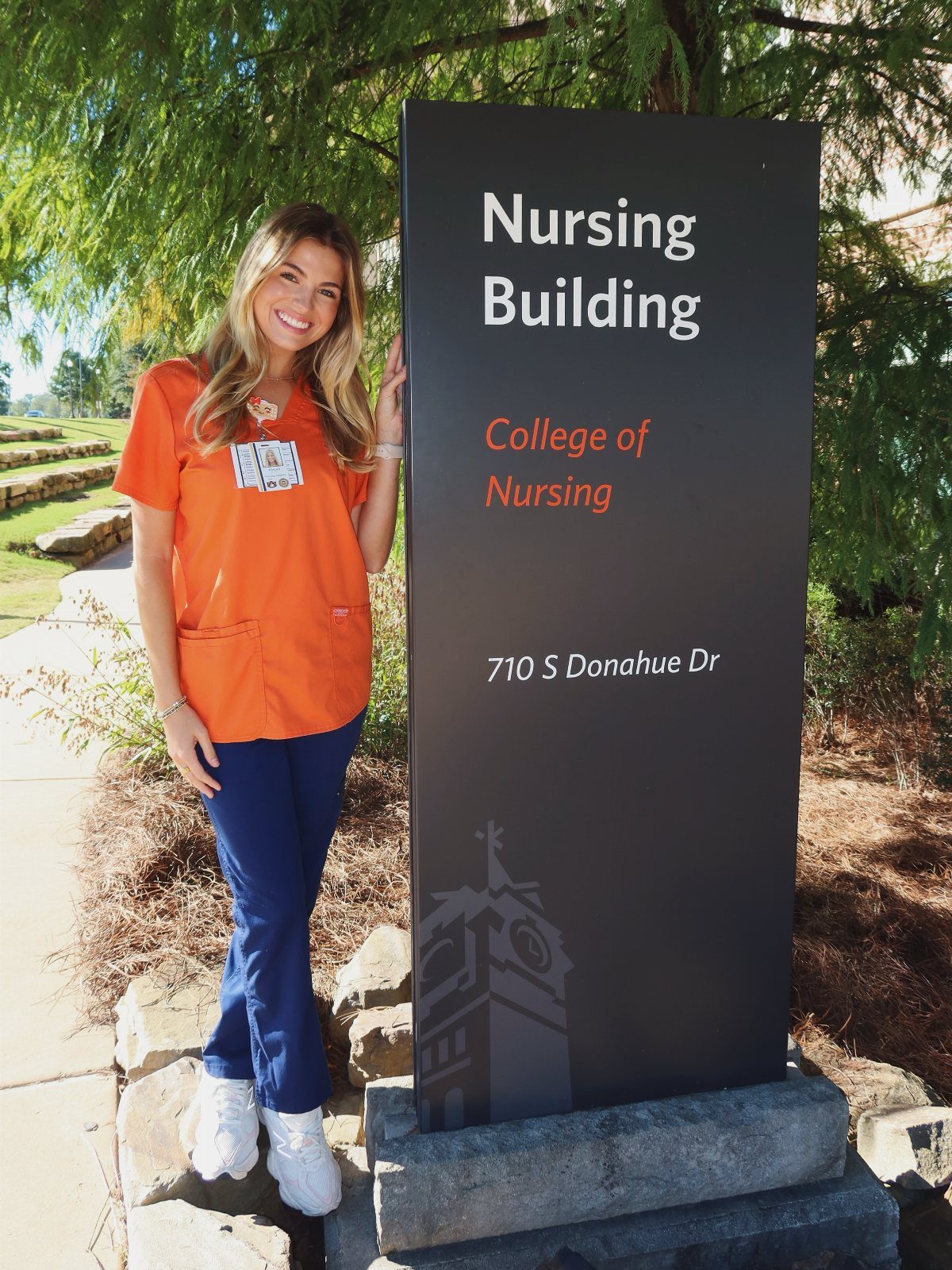 Smiling woman in orange and blue scrubs standing beside a Nursing Building sign.