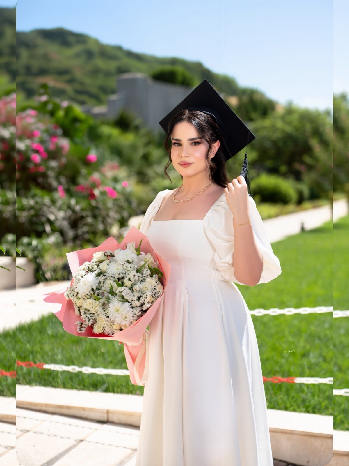 Woman in white dress and graduation cap holding flowers in a sunny garden
