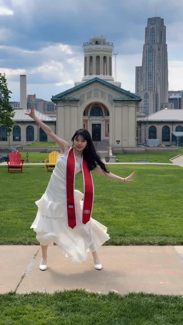 Woman in white dress and red graduation stole posing before a city building.