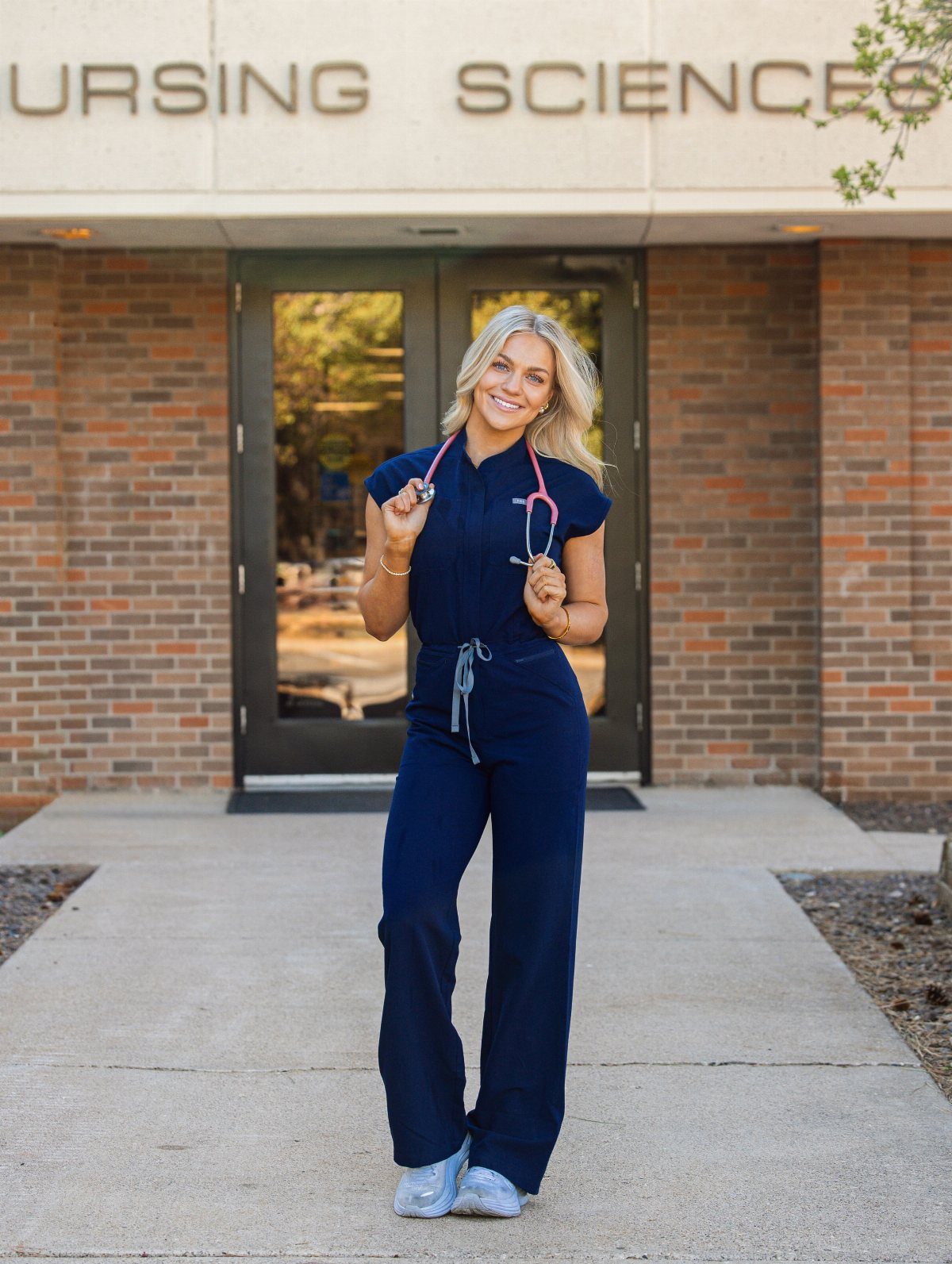 Blonde woman in navy blue scrubs with a pink stethoscope posing outdoors.