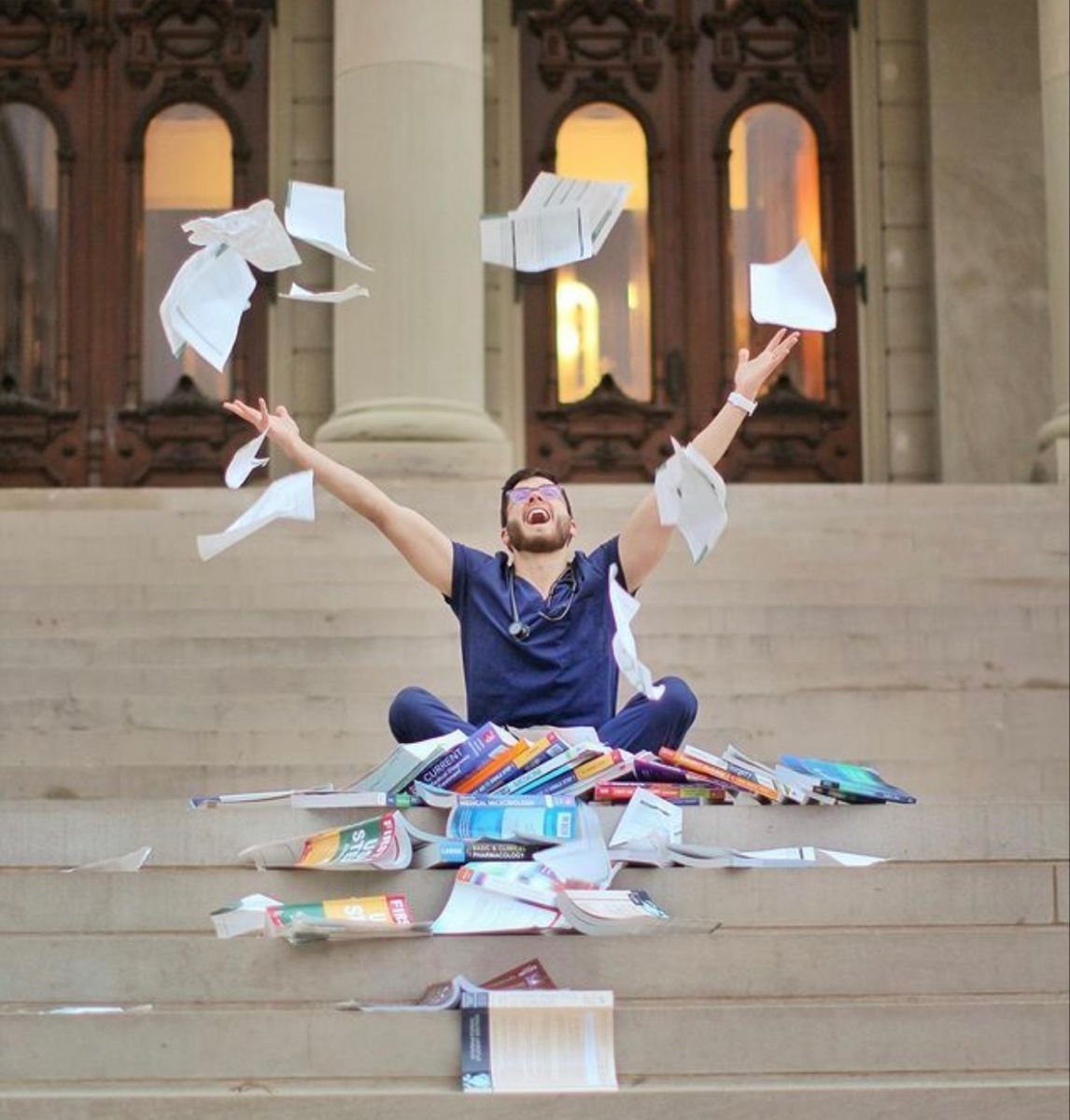 Man in blue scrubs sitting on steps throwing papers in the air celebrating graduation