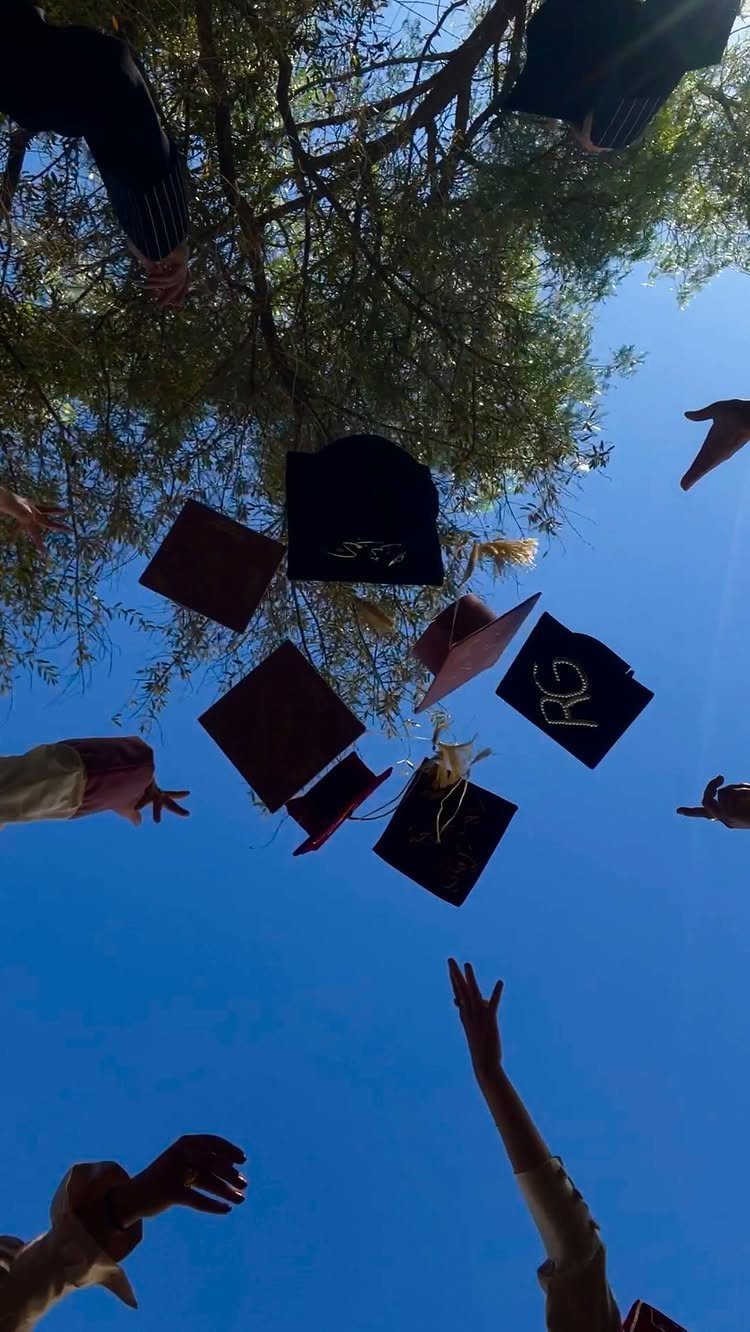 Graduation caps tossed into a blue sky with green tree branches above.
