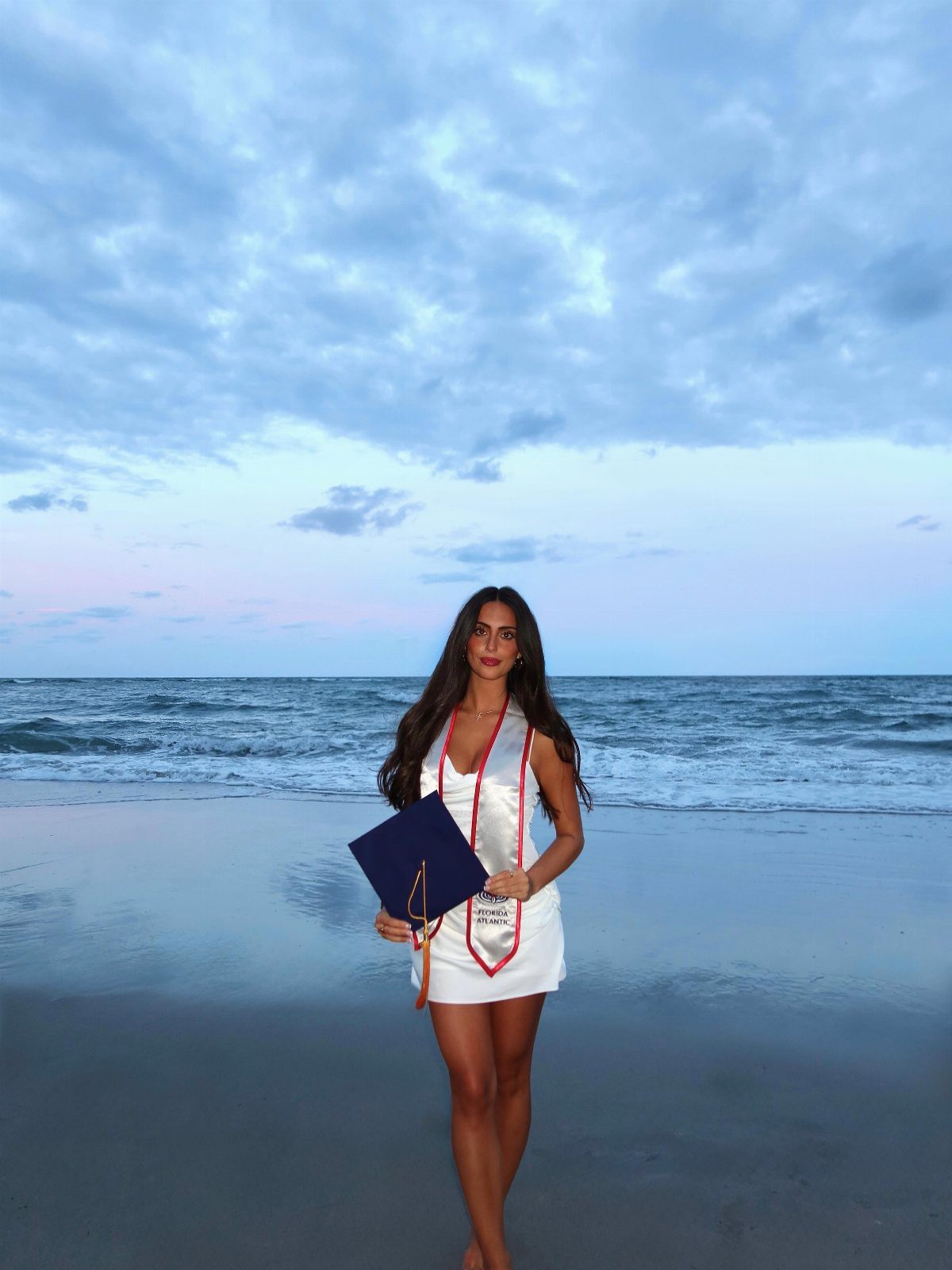 Woman in white dress and graduation stole holding diploma on a sandy beach