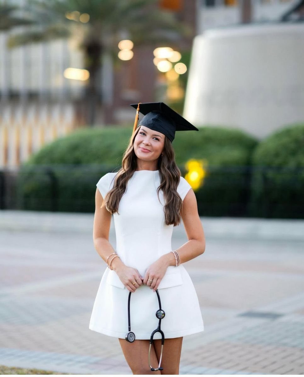 Woman in white dress and graduation cap holding a stethoscope outdoors