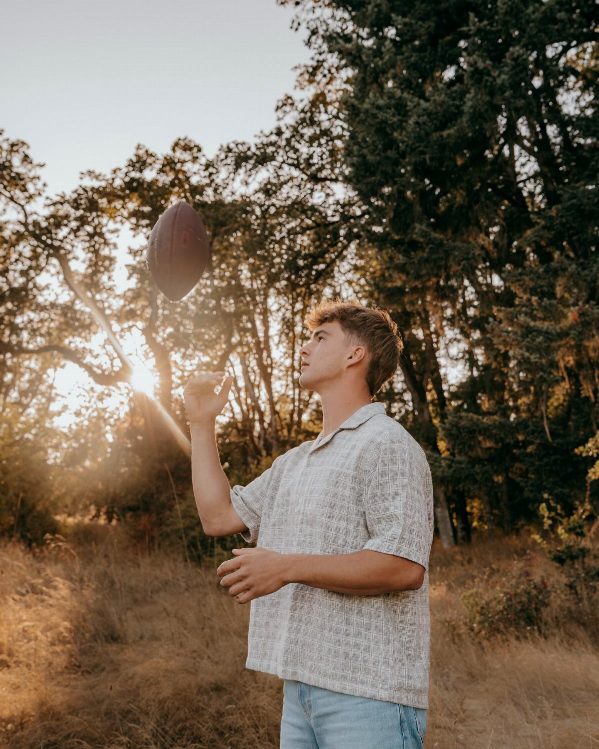 Young man tossing a football in a sunny field with trees behind him