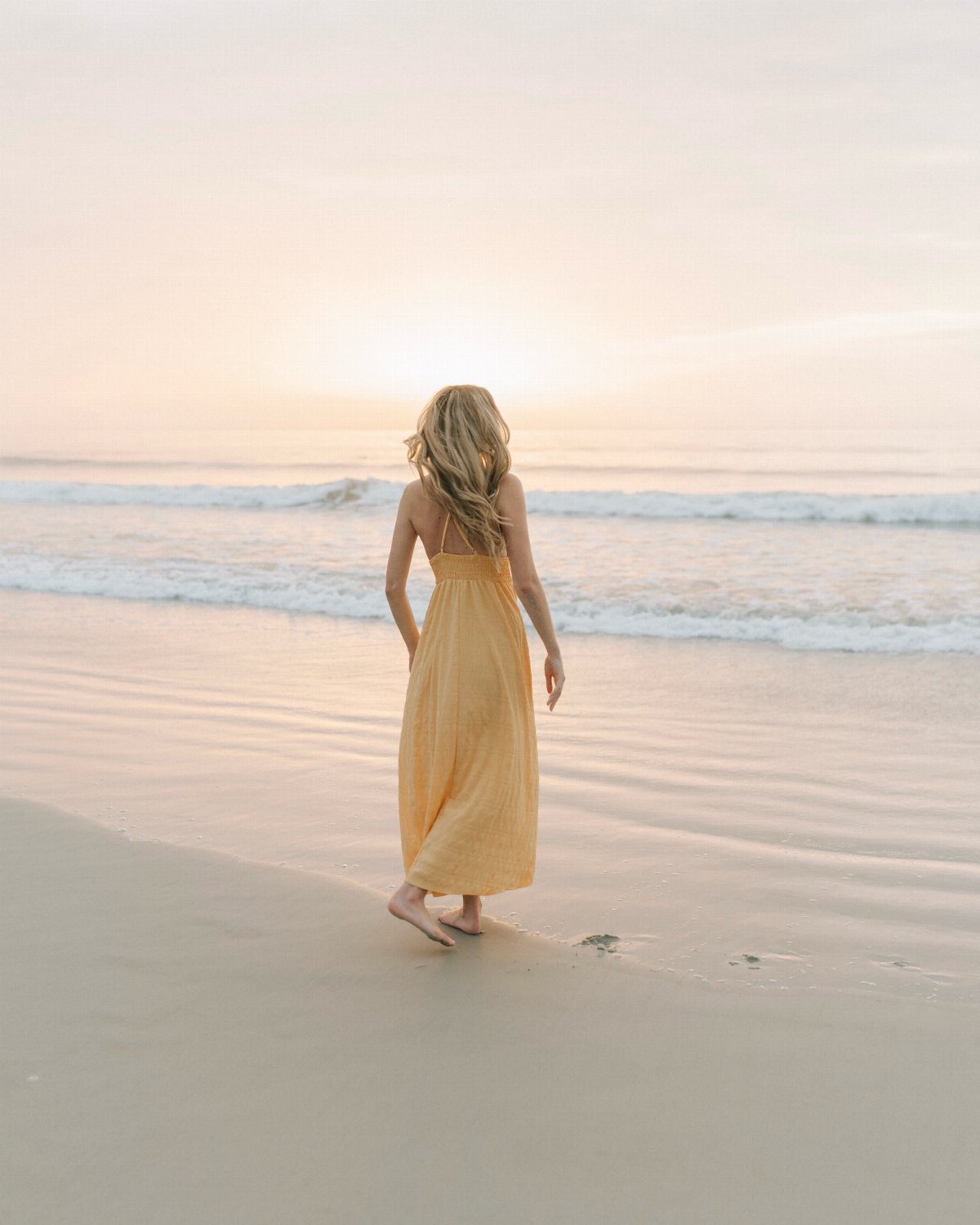 Woman in yellow dress standing on a beach facing the setting sun.