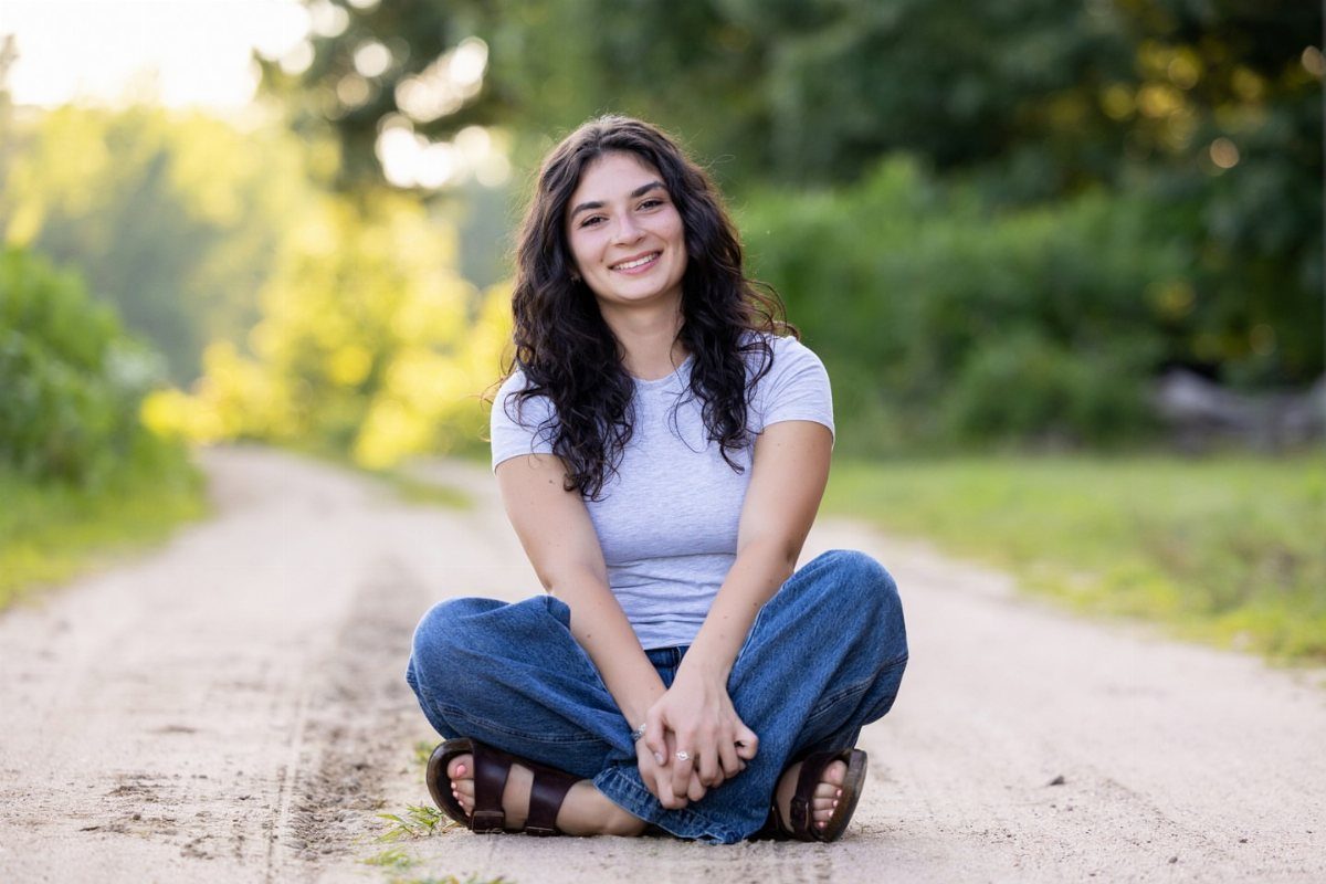 Young woman sitting cross-legged on a dirt road surrounded by blurred green trees.