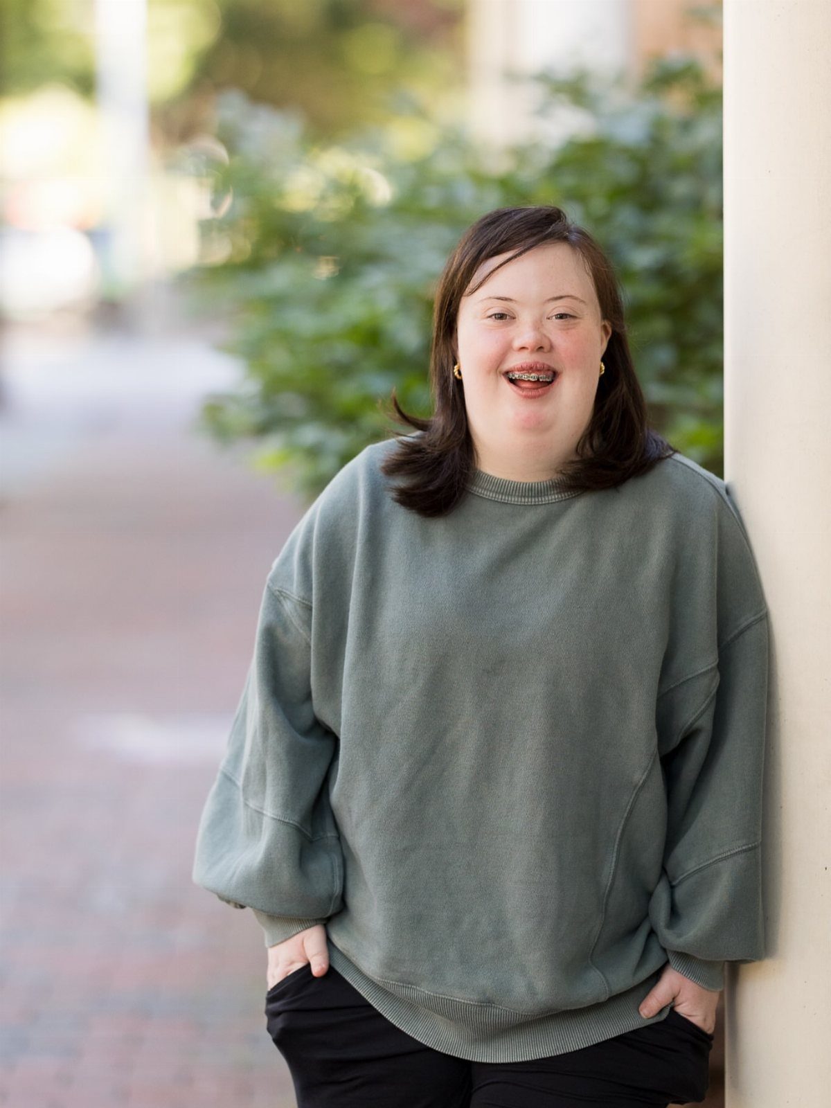 Smiling young woman in a green sweater leaning against a white wall outdoors.