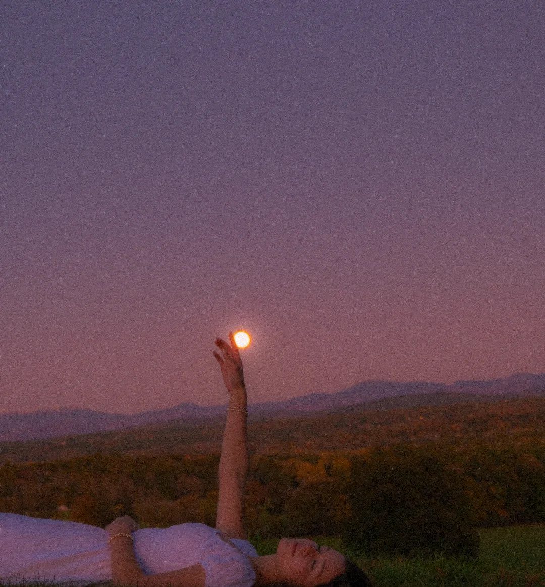 Person lying in a field holding the setting sun in their open palm.