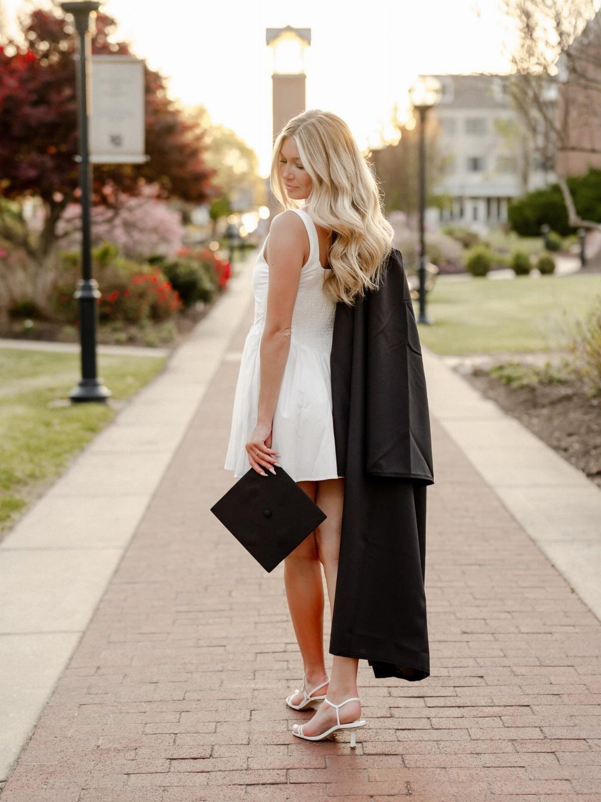 Blonde woman in white dress holding a graduation cap and gown on campus