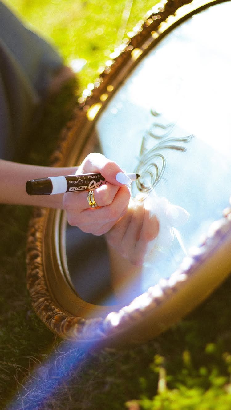 Hand writing with a black marker on a gold-framed mirror on green grass.