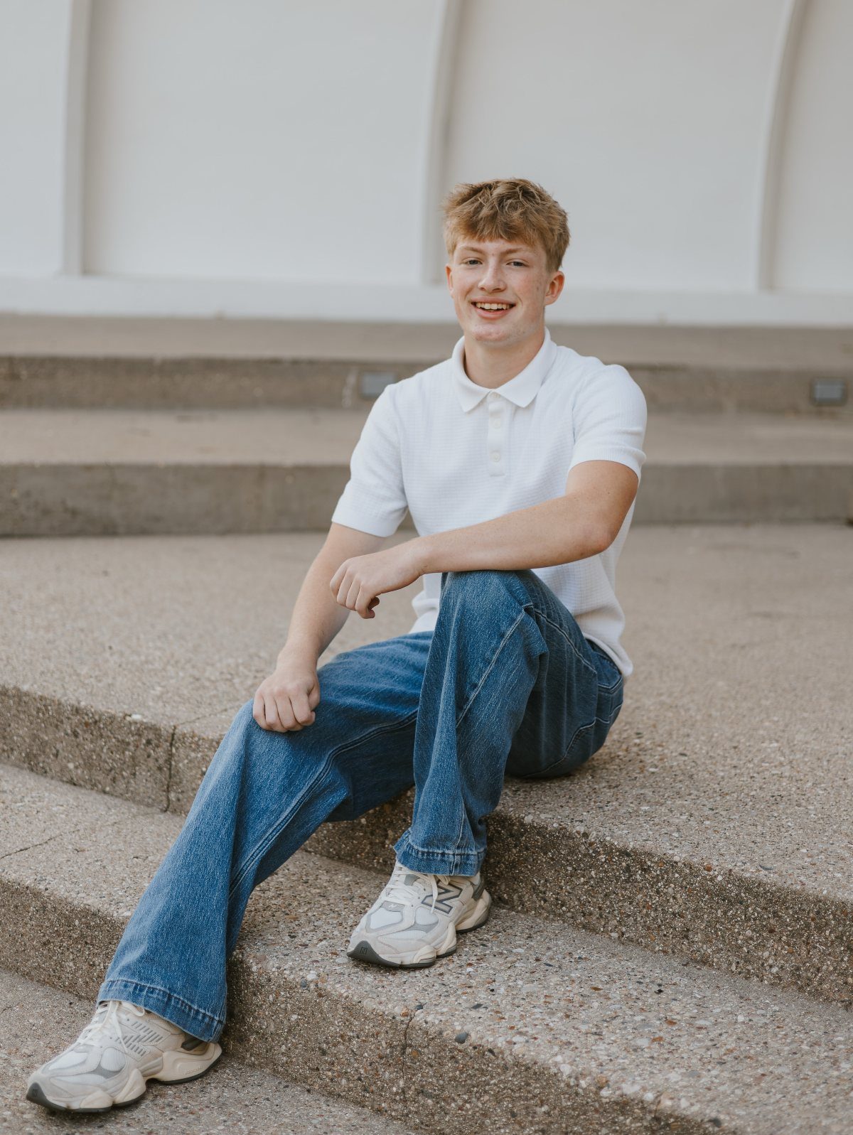 Young man in white polo and jeans sitting on concrete steps against white wall