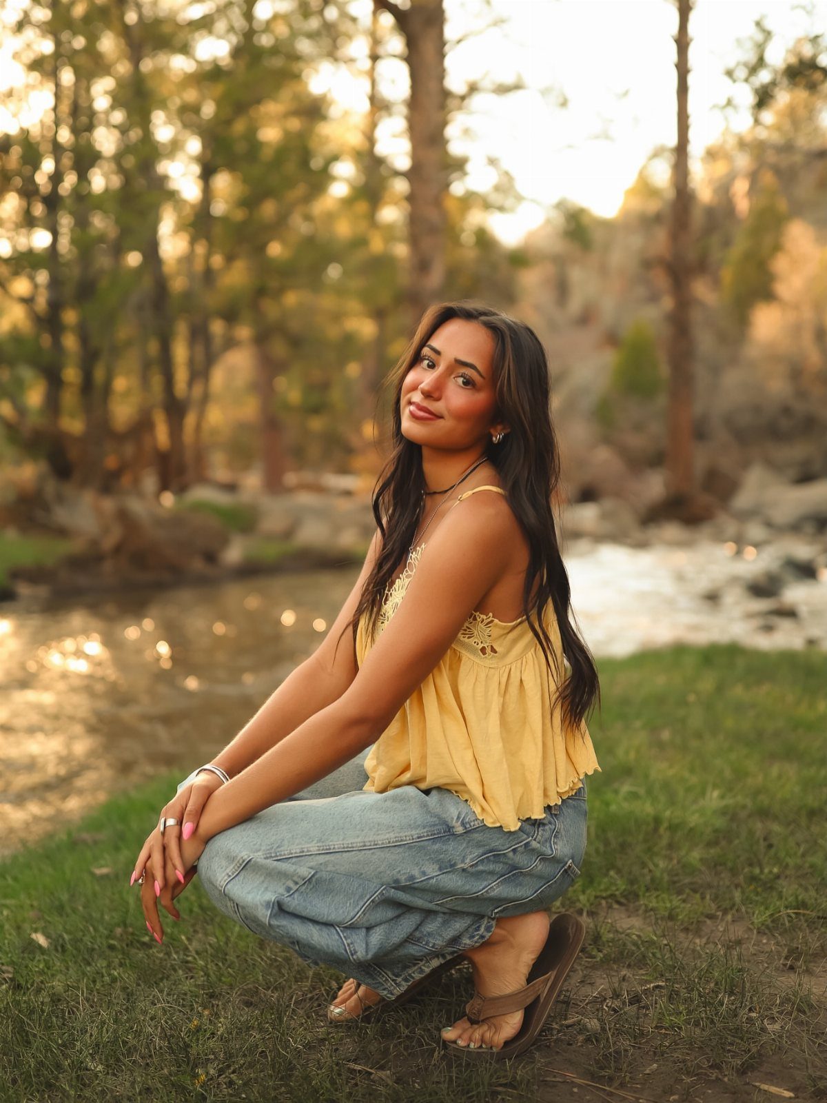 Young woman in yellow top and jeans crouching by a river in woods.