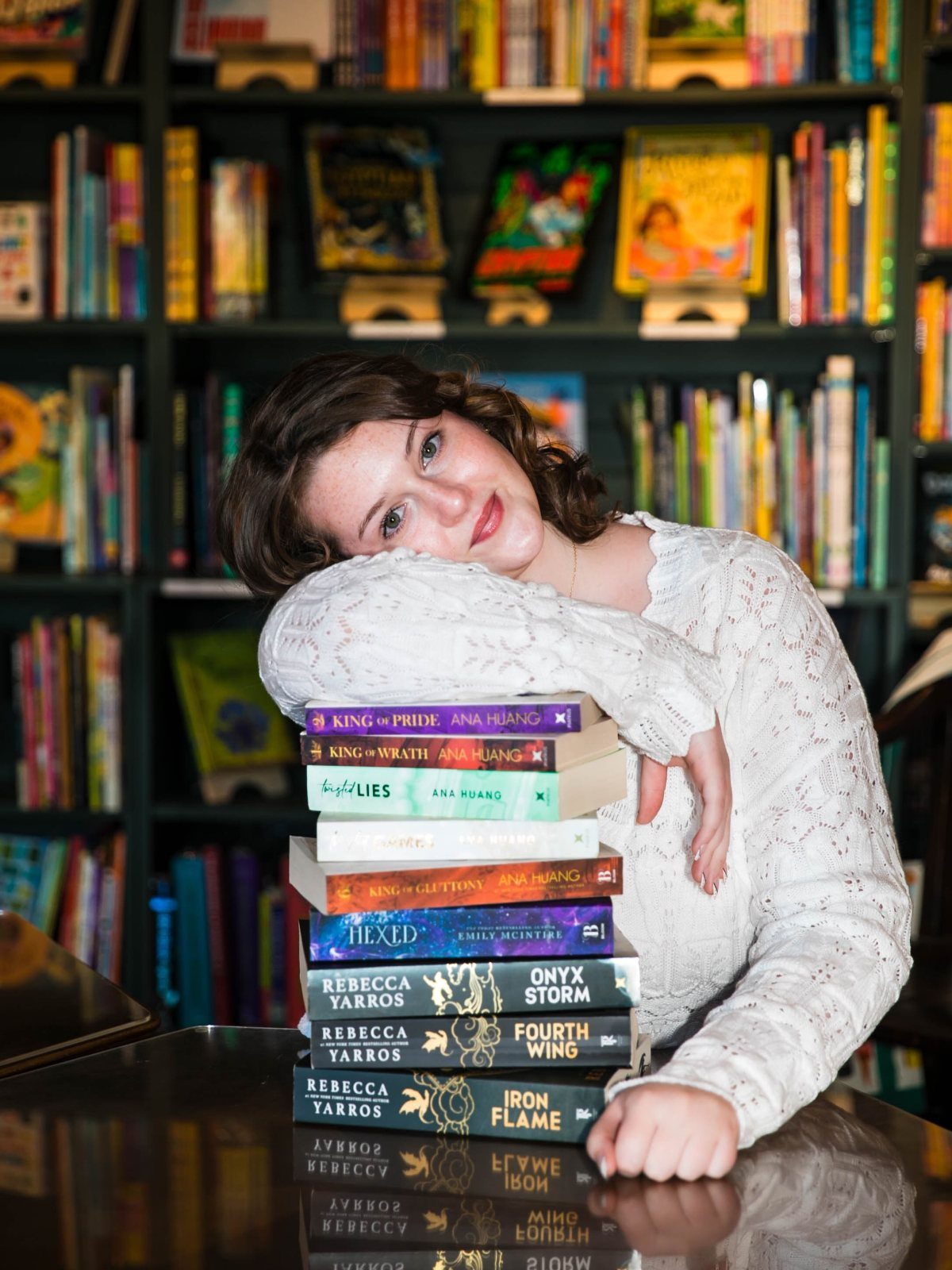 Young woman leaning on a stack of books in front of bookshelves.