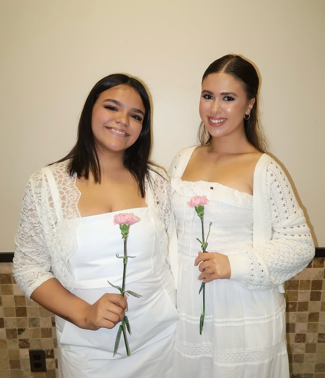 Two women in white dresses holding pink carnations and smiling for the camera.