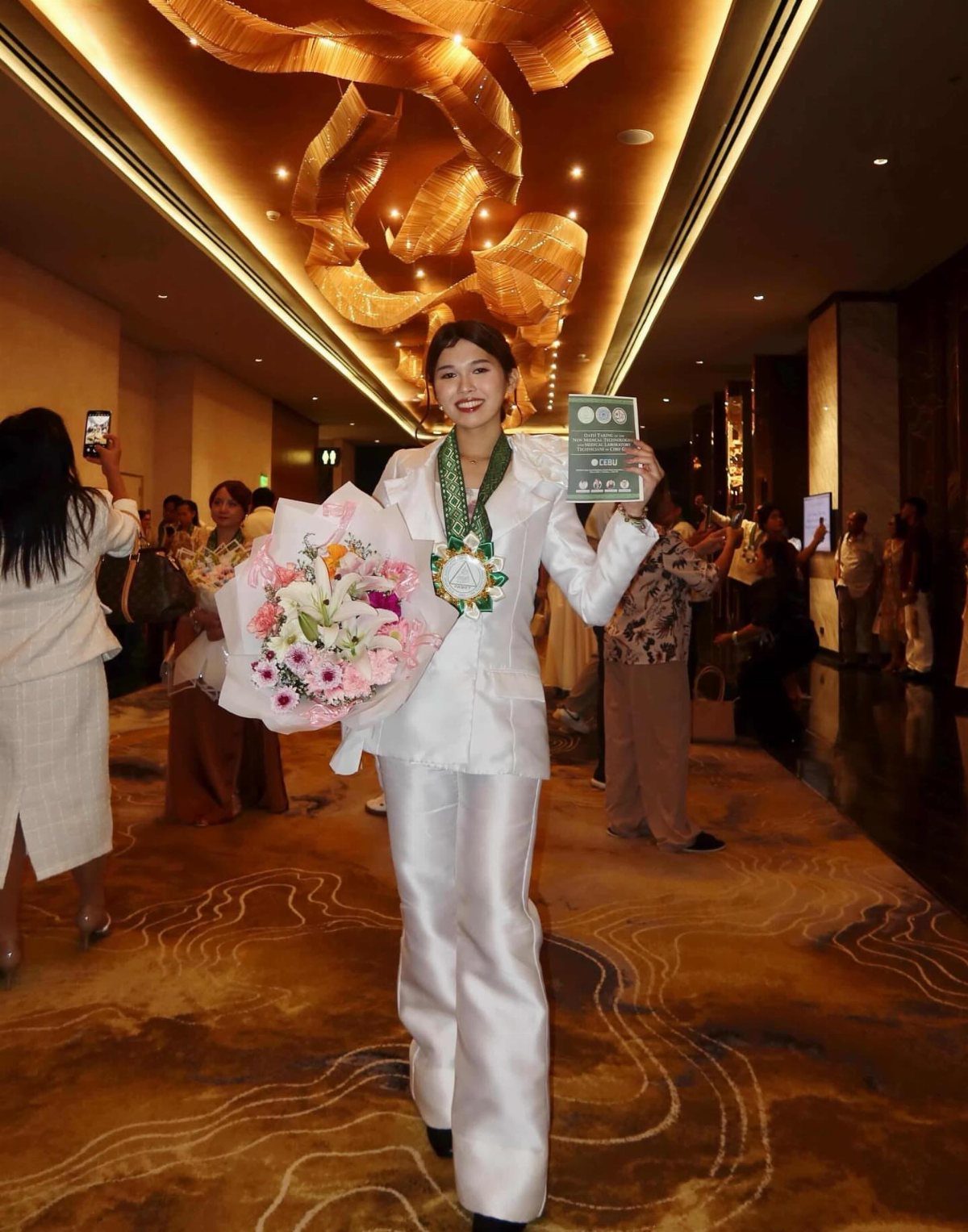 Woman in white satin pant suit holding flowers and a certificate in hallway