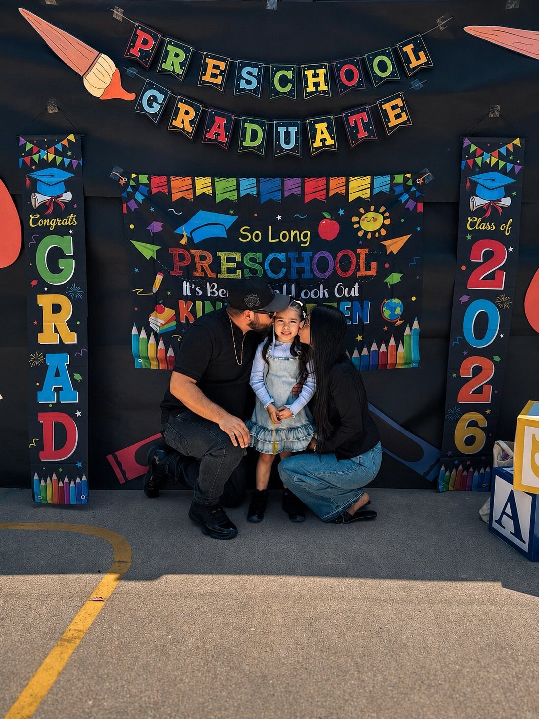 Young girl in blue dress being kissed by parents before preschool graduation backdrop