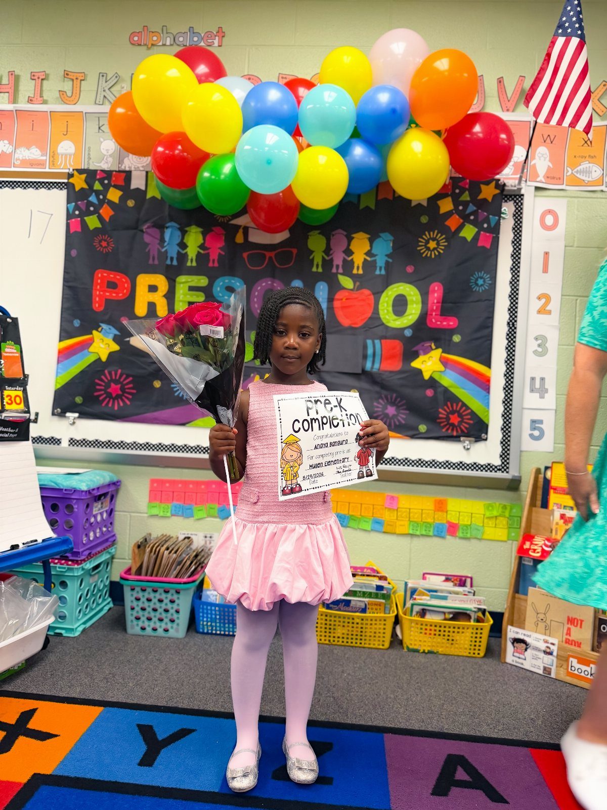 Young girl in pink dress holding flowers and certificate in a preschool classroom