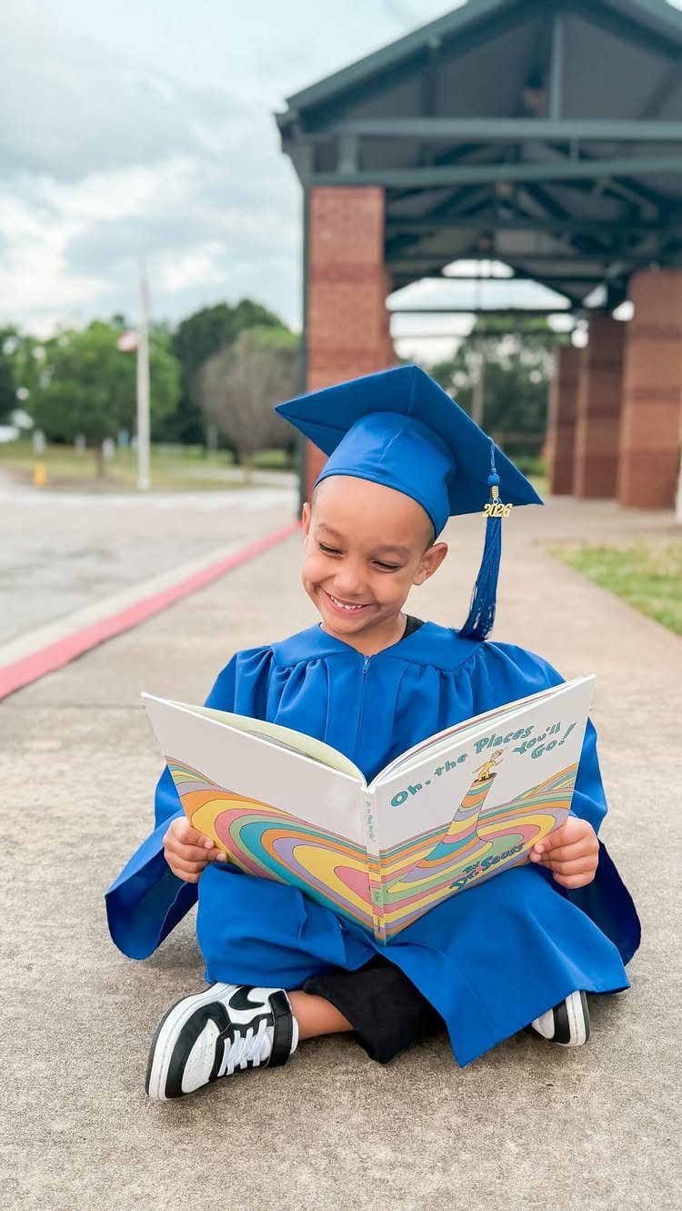 Young child in blue graduation cap and gown reading a colorful book outdoors.