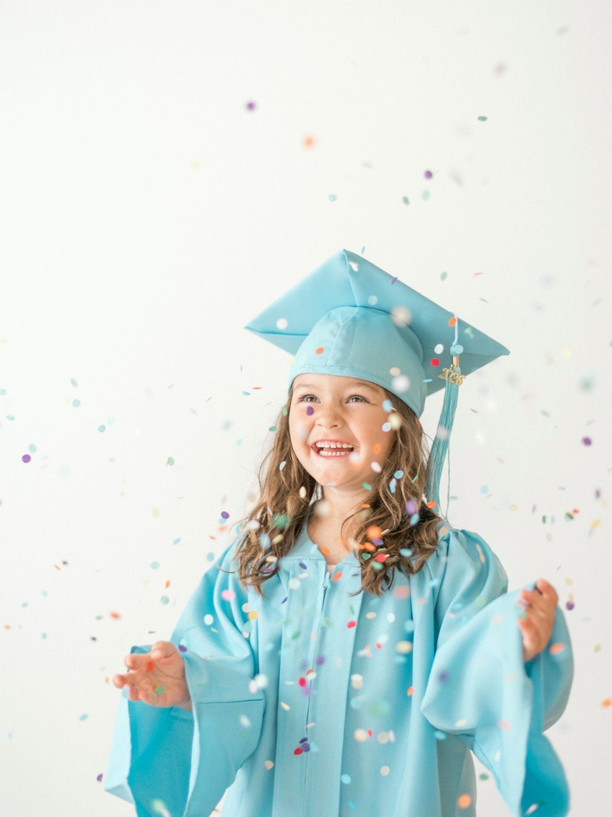 Young girl in light blue graduation cap and gown smiling amidst falling confetti
