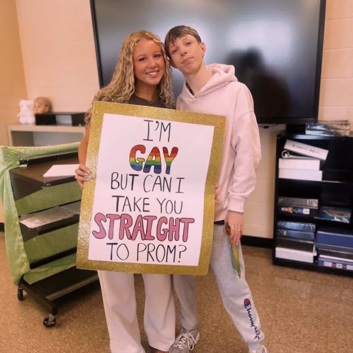 Two teenagers holding a rainbow themed promposal poster in a classroom setting.