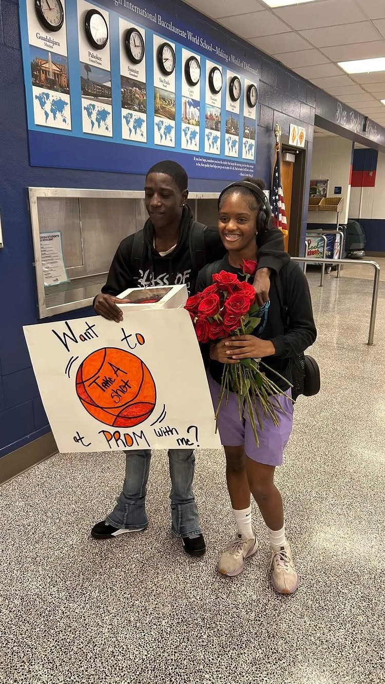 Teenage couple in a school hallway with a basketball promposal poster and roses.
