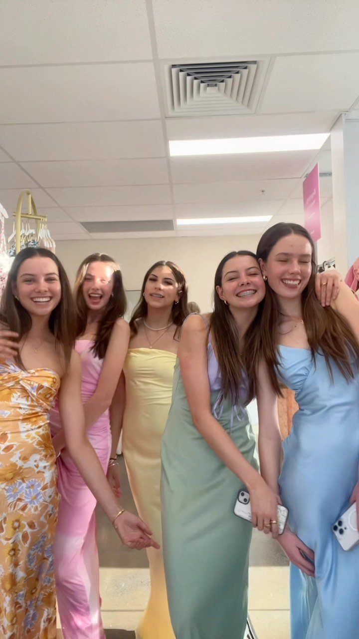 Five young women in colorful formal gowns posing together in a dress shop.