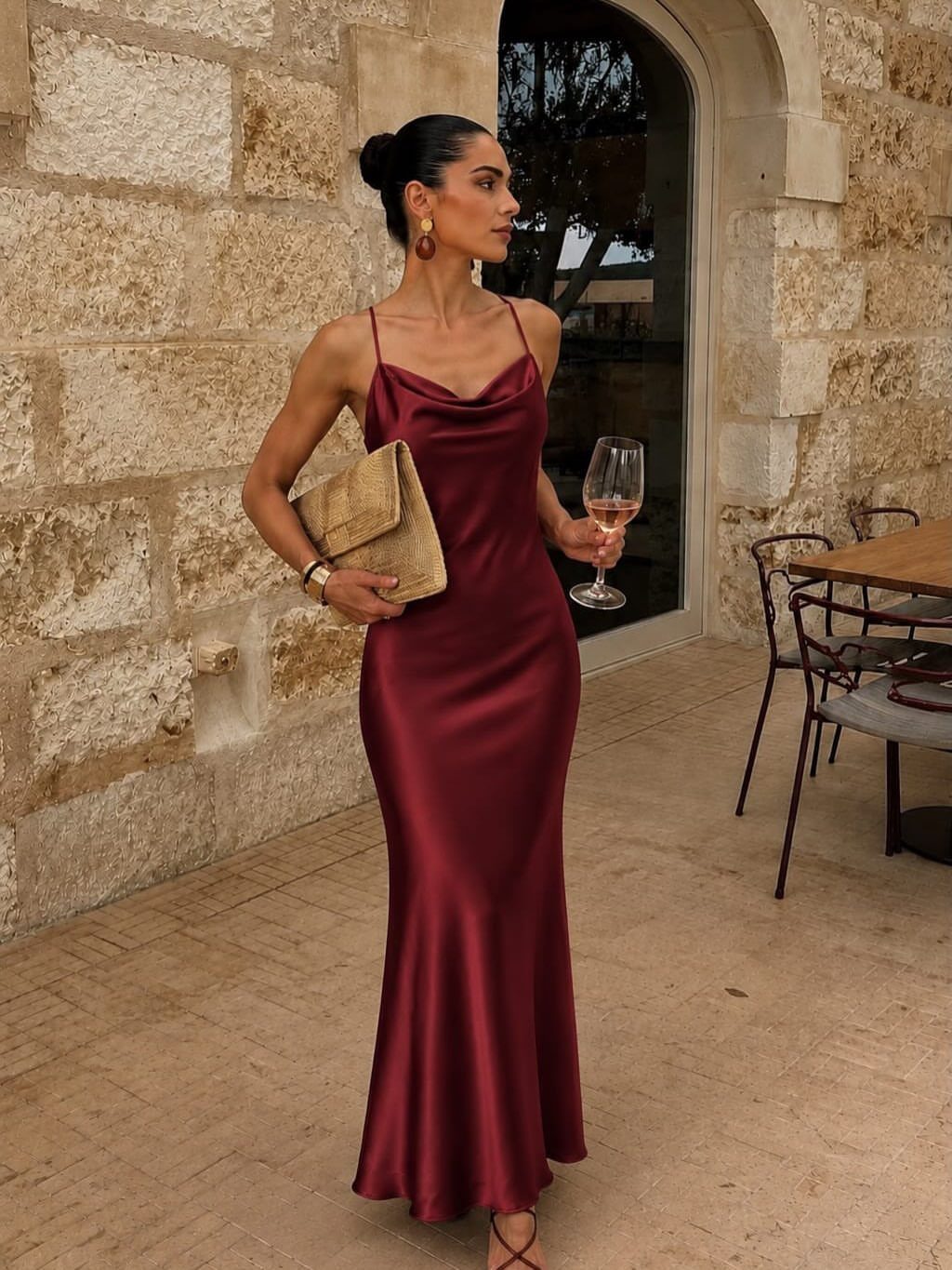 Woman in a burgundy satin cowl neck maxi dress holding a gold clutch.