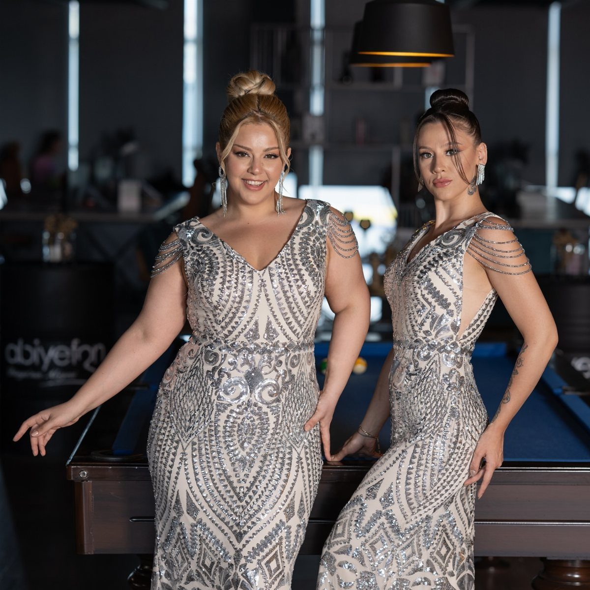 Two women in silver sequined gowns posing next to a blue pool table.