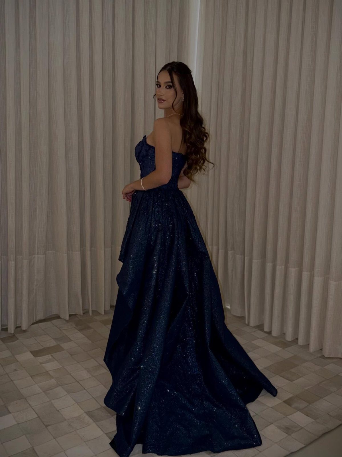 Woman in a strapless navy blue glittery floor-length gown posing against white curtains.
