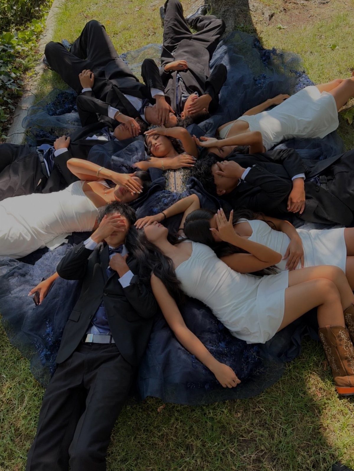 Group of teenagers in formal wear lying in a circle on green grass.