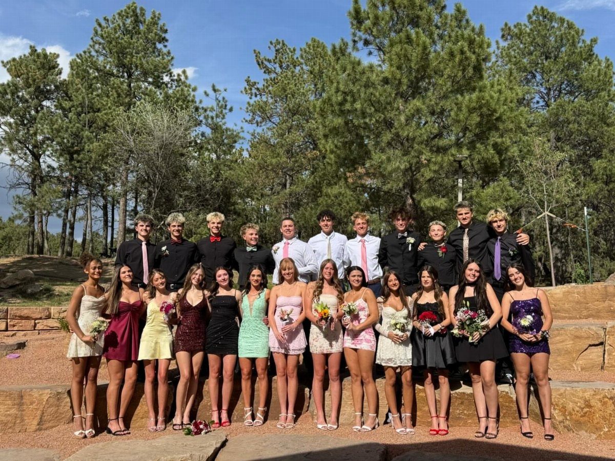 Group of teenagers in formal prom dresses and suits posing outdoors by trees.