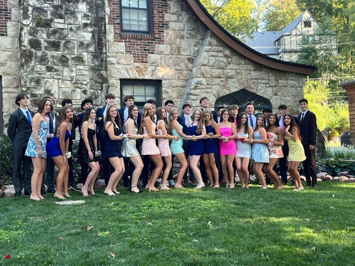 Teenagers in formal prom attire posing in a line on a green lawn.