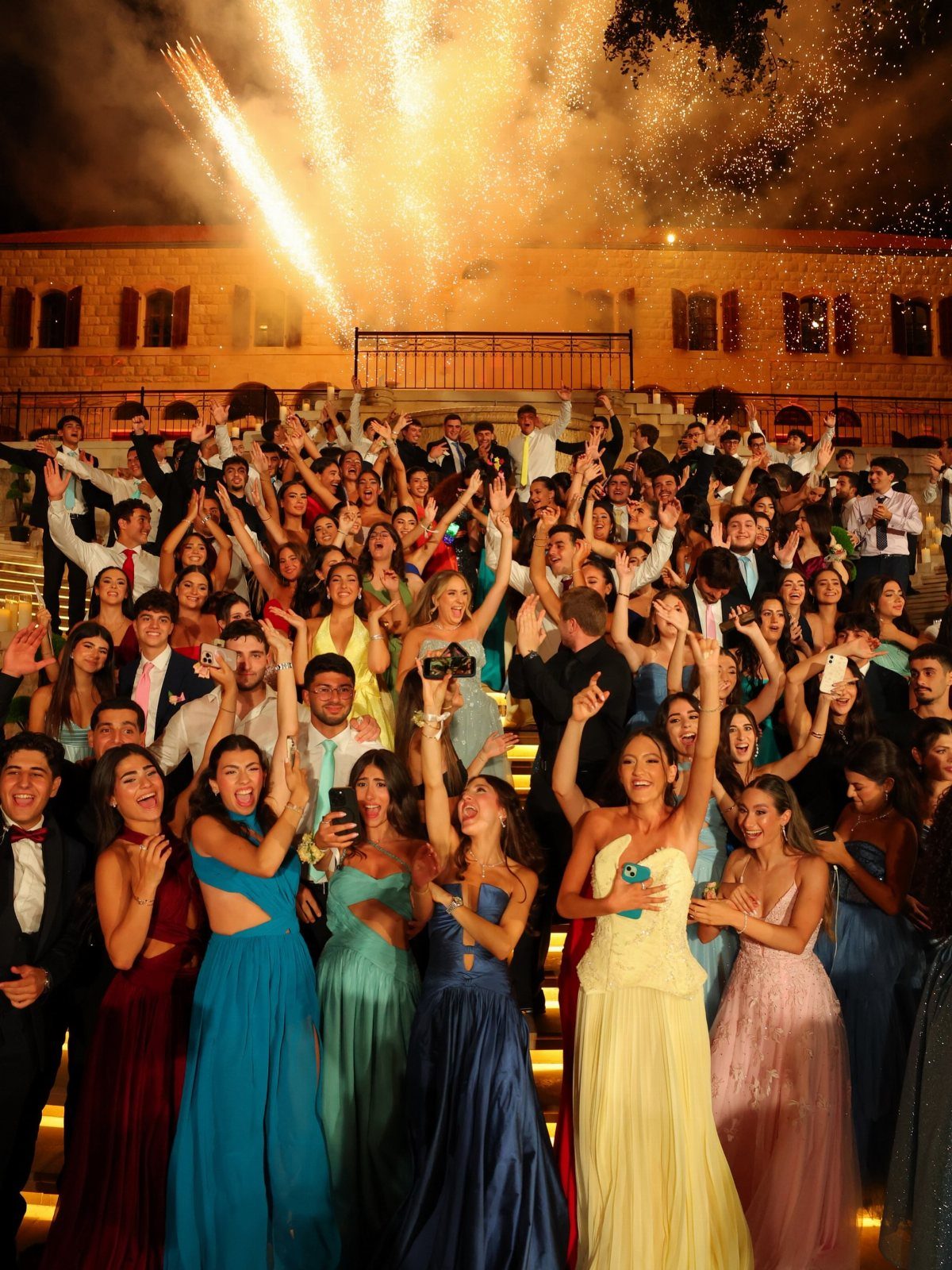 Group of students in formal wear posing on stairs with fireworks overhead.