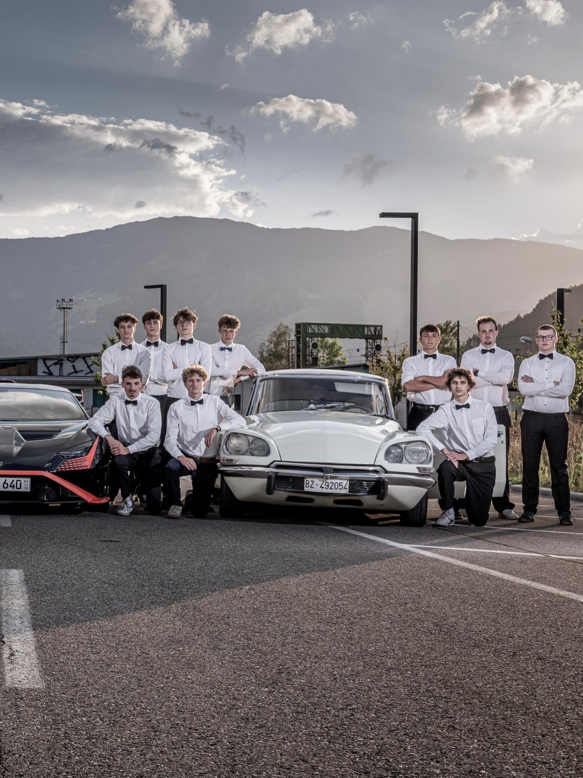 Group of young men in formal wear posing with vintage and modern cars.