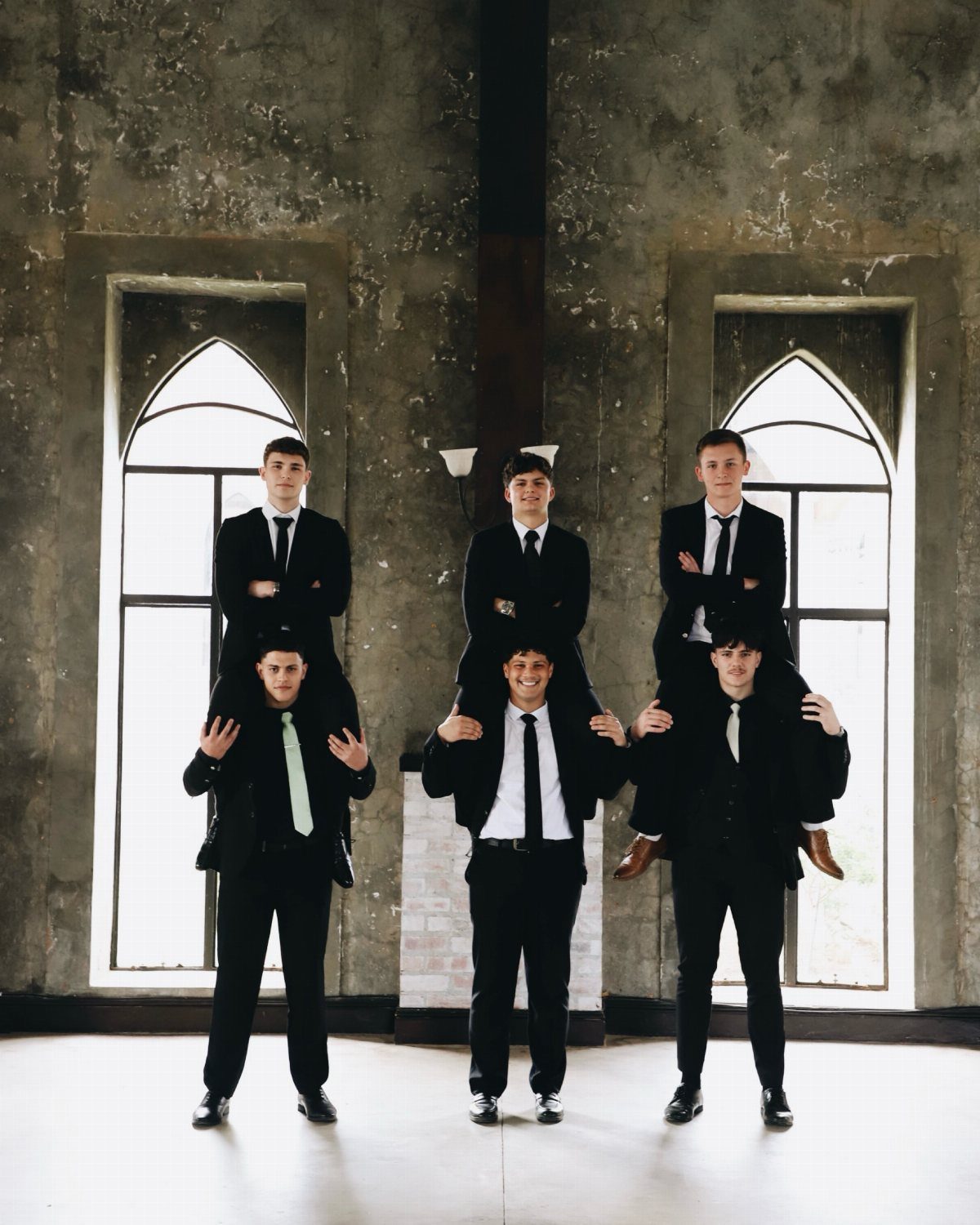 Six young men in black suits posing in a two-tier group formation indoors.