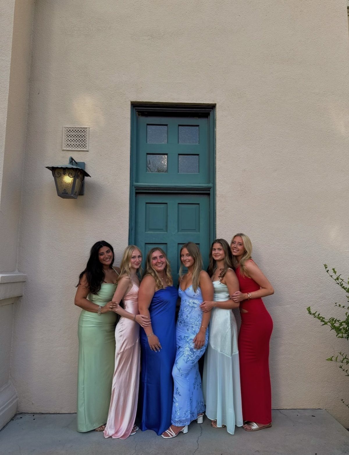 Six young women in colorful formal gowns posing together in front of a door.