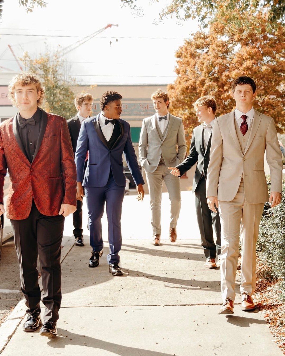 Six young men in formal suits walking together on a sunny outdoor sidewalk.