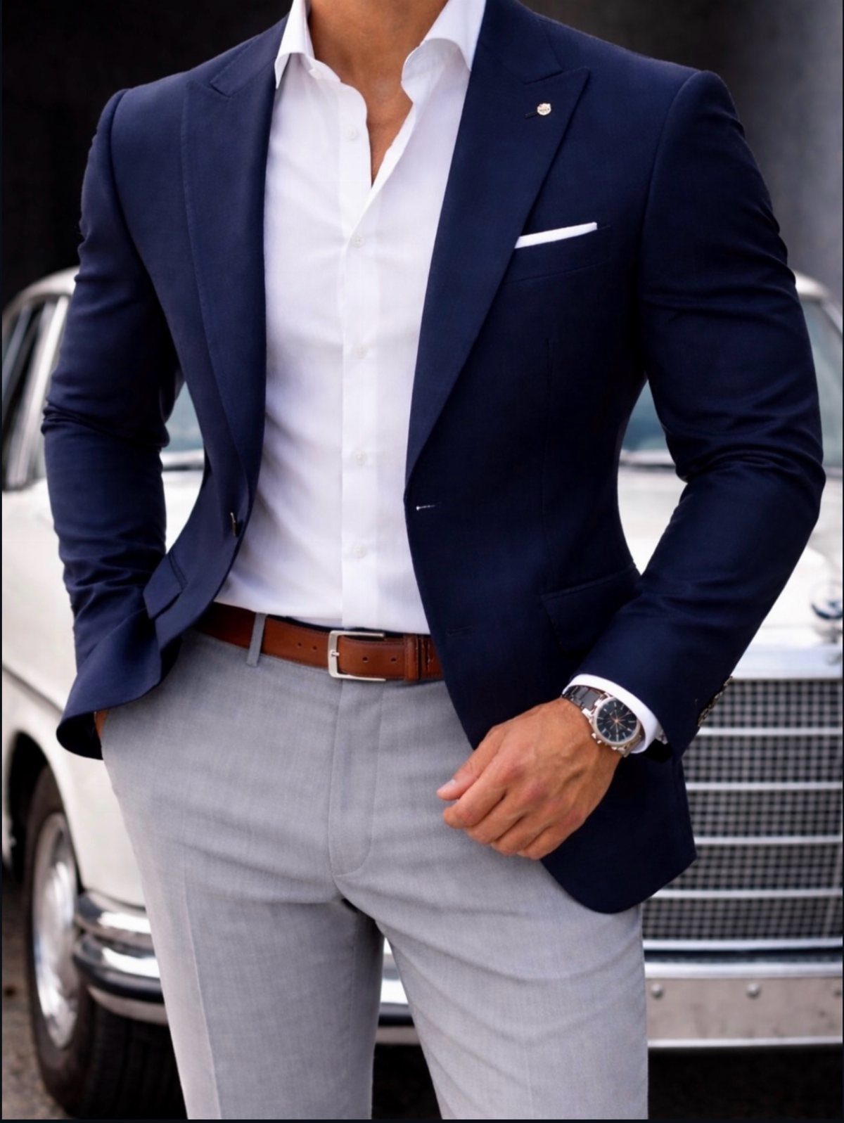 Man wearing a navy blue blazer, white shirt, and grey trousers by a car.