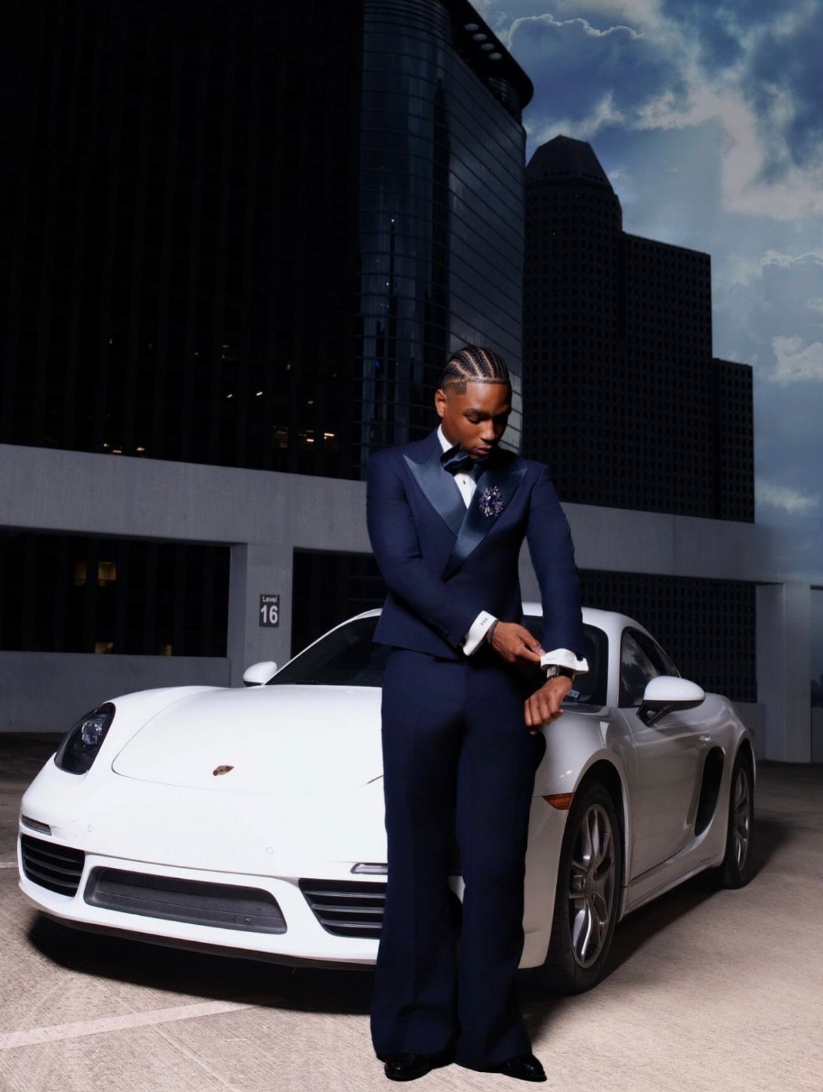 Young man in a navy blue tuxedo standing beside a white sports car.