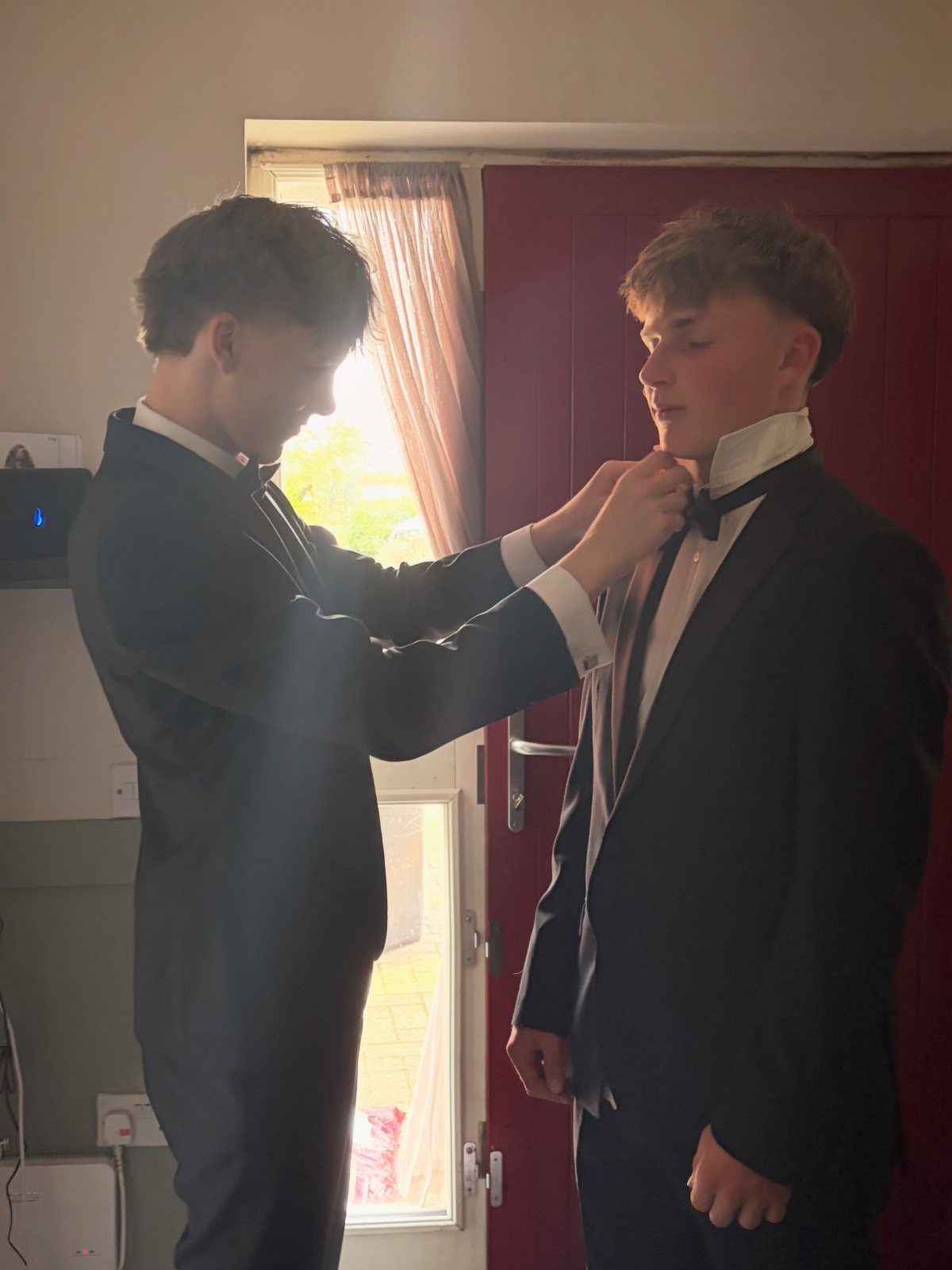 Two young men in tuxedos, one adjusting the other's bowtie indoors.